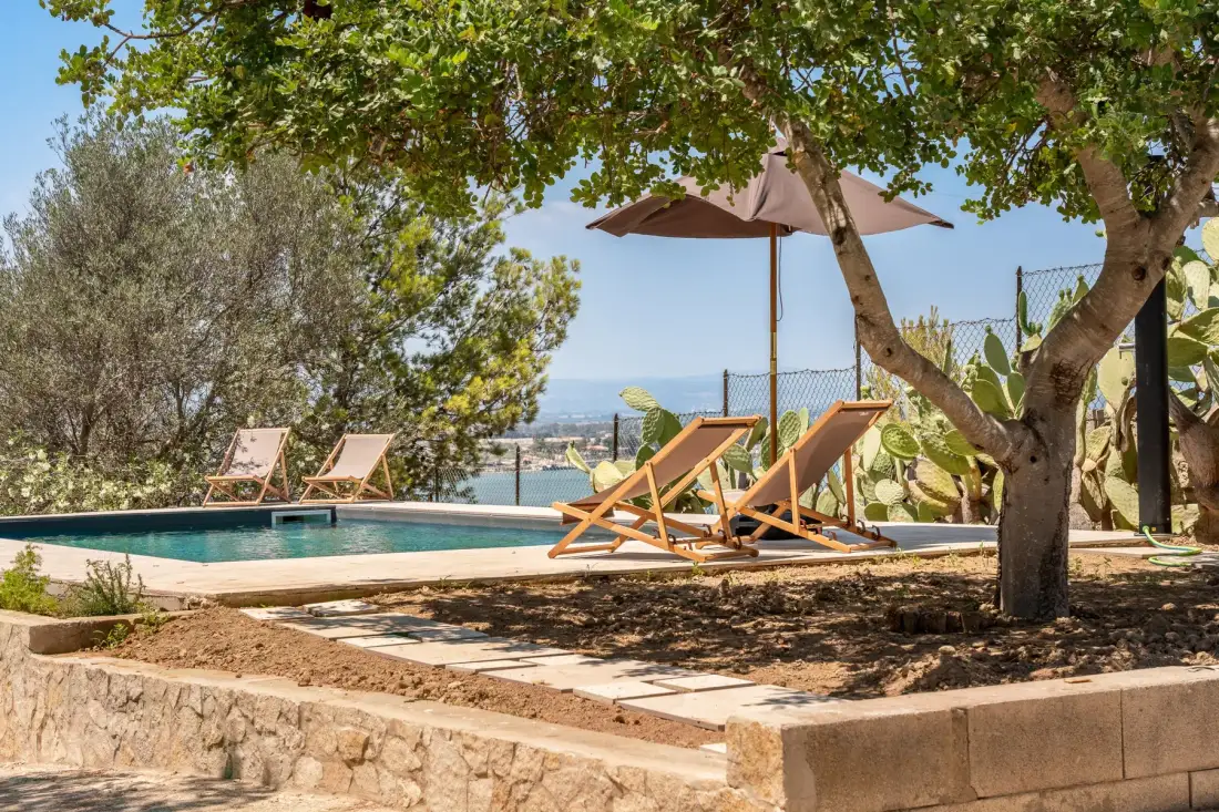 A swimming pool with lounge seating and an umbrella sits beneath leafy trees.