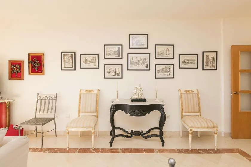 A bright room with framed artwork above a decorative console table and several seats.