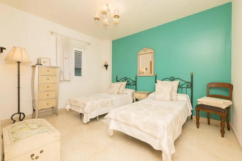 A bedroom with two beds, a turquoise accent wall, and a small chair.