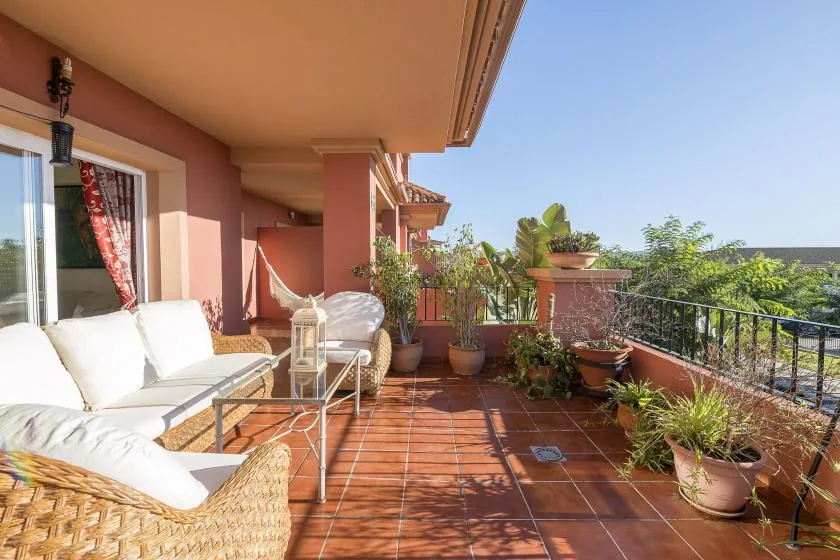 A sunlit terrace with outdoor seating, potted plants, and a metal railing.