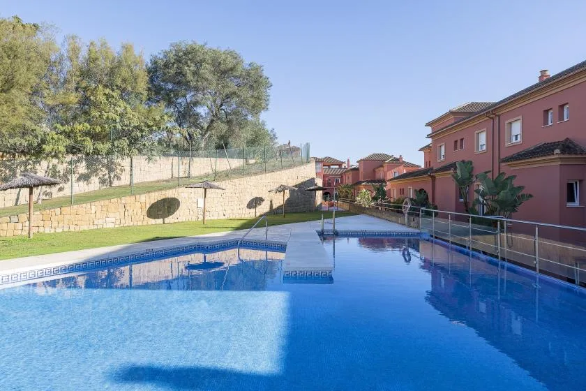 An outdoor swimming pool beside red residential buildings, with trees along the wall.