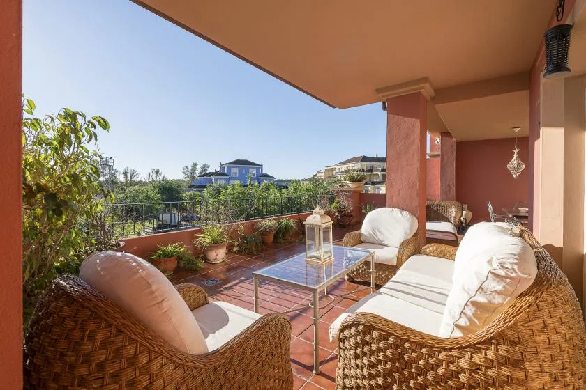 A covered terrace with wicker seating, a glass table, and potted plants.