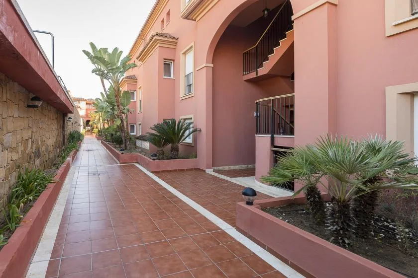 A tiled courtyard walkway runs beside a pink building and planted garden beds.
