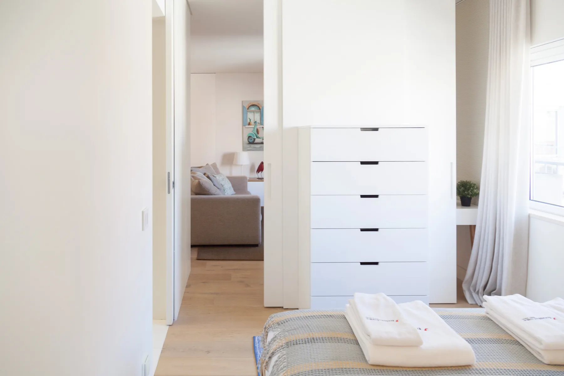 Un dormitorio luminoso con una cómoda blanca, toallas dobladas y una vista a una zona de estar.
