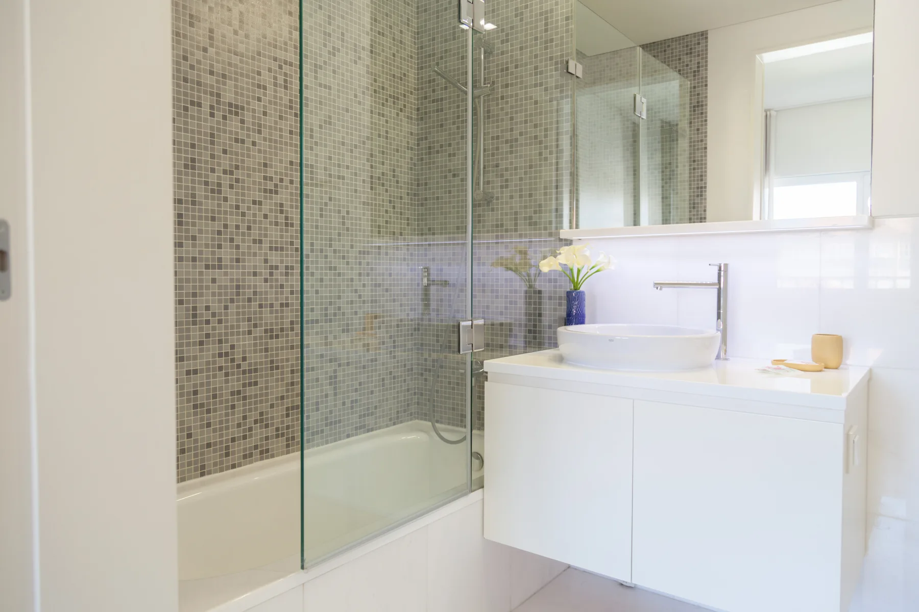 Un baño luminoso con bañera tras una mampara de vidrio y mueble blanco.