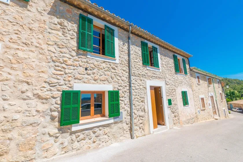 A stone building with green shutters stands beside a sunny paved lane.