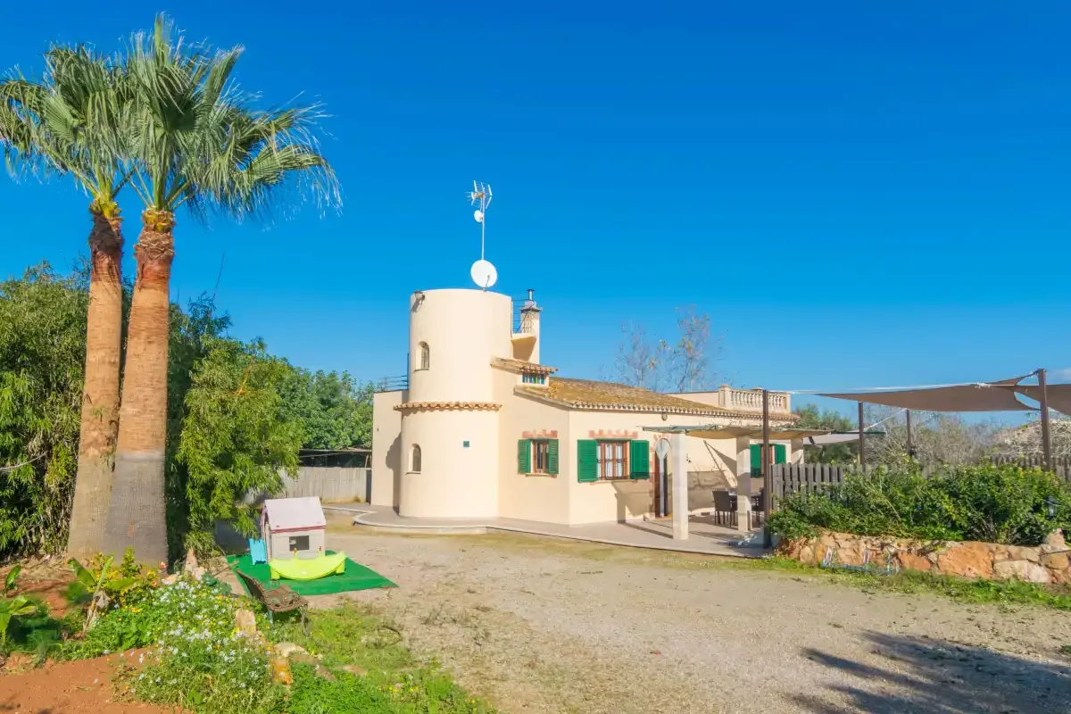 A white villa sits beside a gravel driveway, with palm trees and a shaded patio.