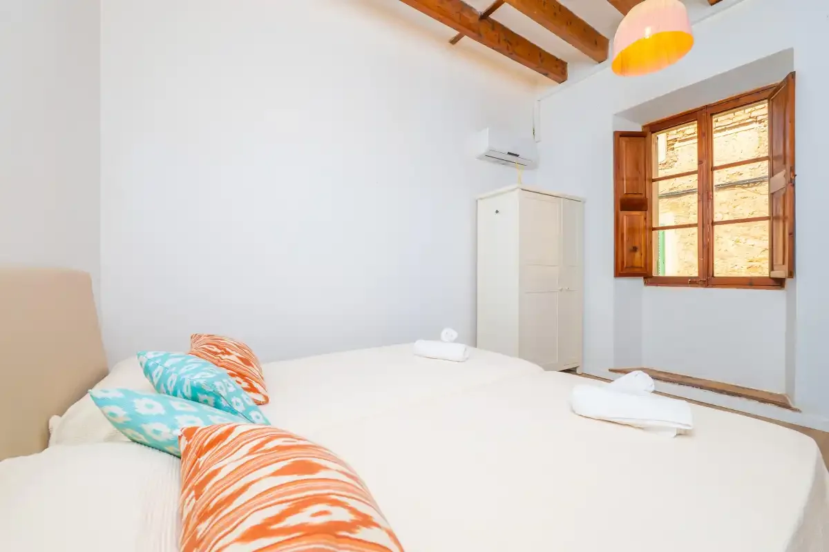 A bedroom with white bedding, colorful pillows, and a wooden window.