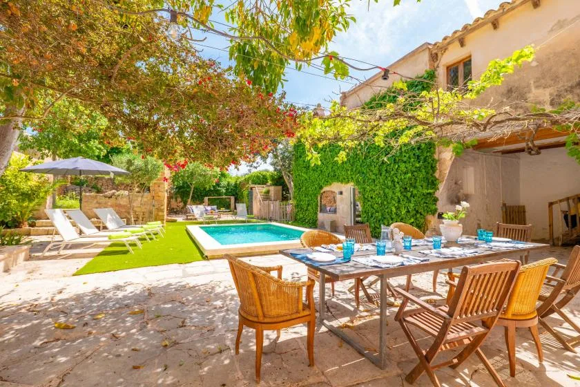 An outdoor dining area overlooks a swimming pool, with lounge seating beneath leafy trees.