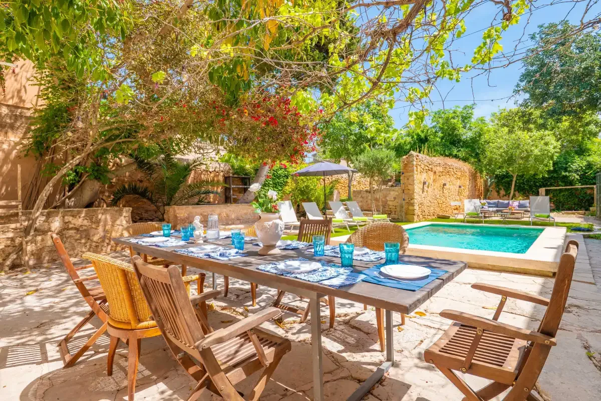 An outdoor dining area beside a swimming pool, shaded by leafy trees.