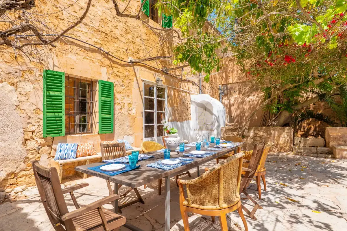 An outdoor dining area beside a stone house, with a table set under leafy branches.