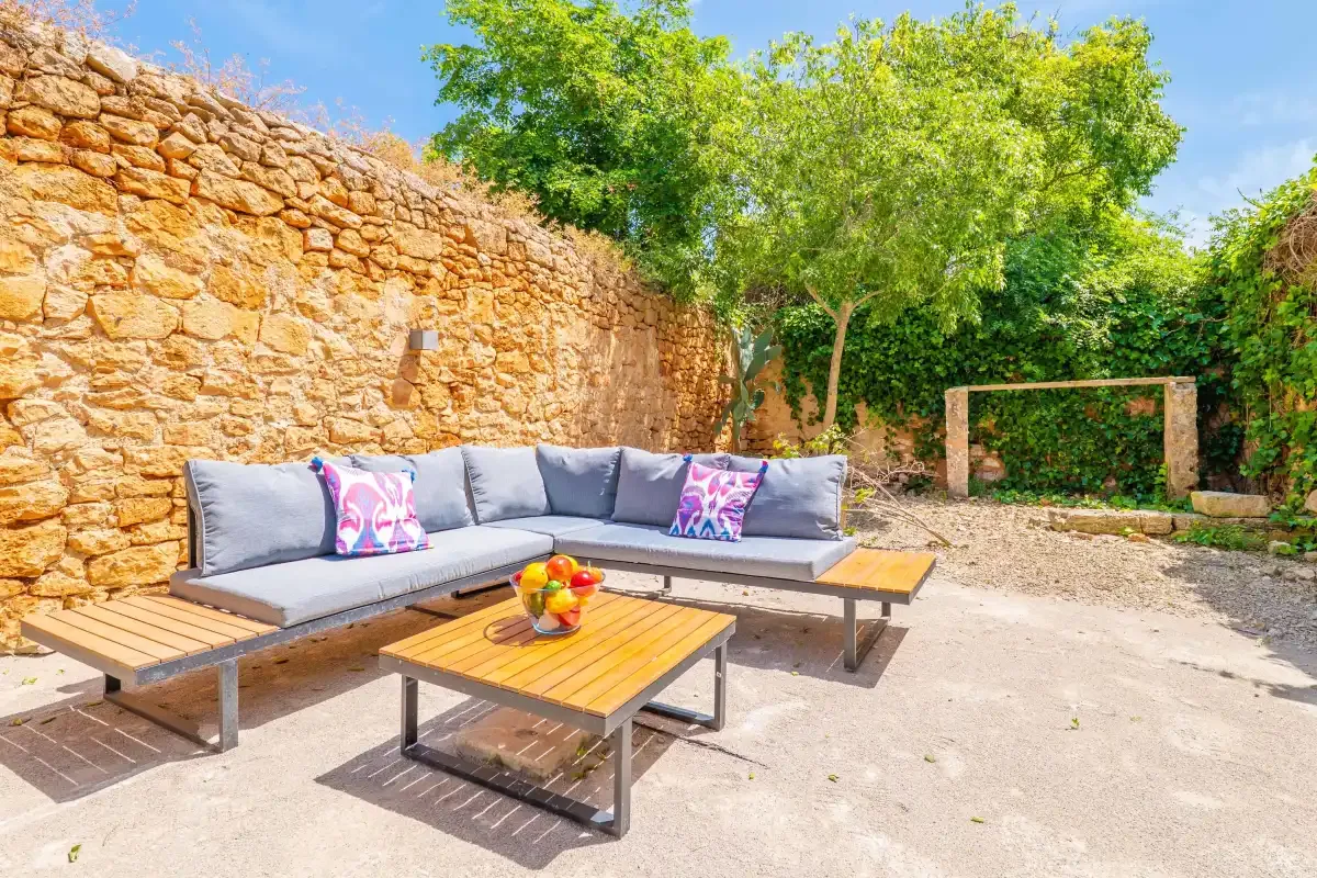 An outdoor seating area with low wooden tables sits beside a stone wall and leafy trees.