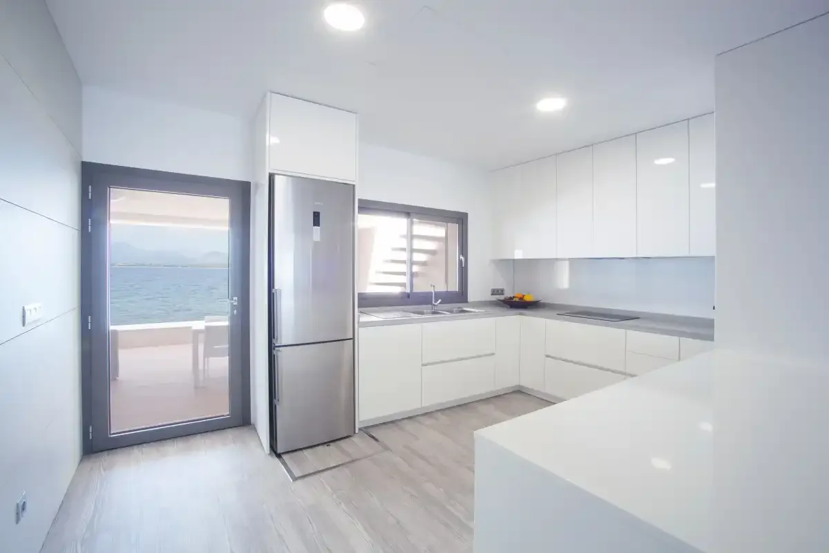 A bright modern kitchen with white cabinets and a view of the sea through the glass door.