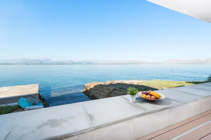 A waterfront patio overlooks calm blue water, with a plate of fruit on the ledge.