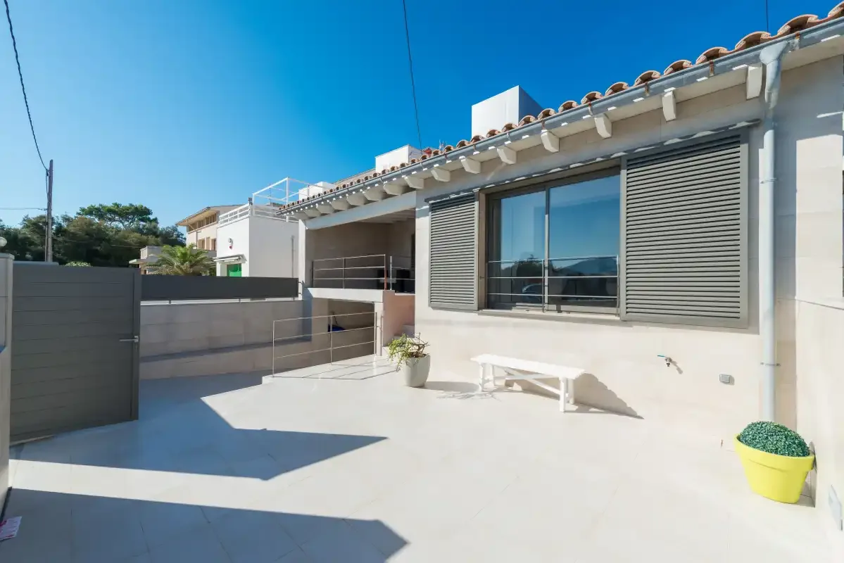 A sunny paved patio beside a modern house, with a large window and outdoor seating.