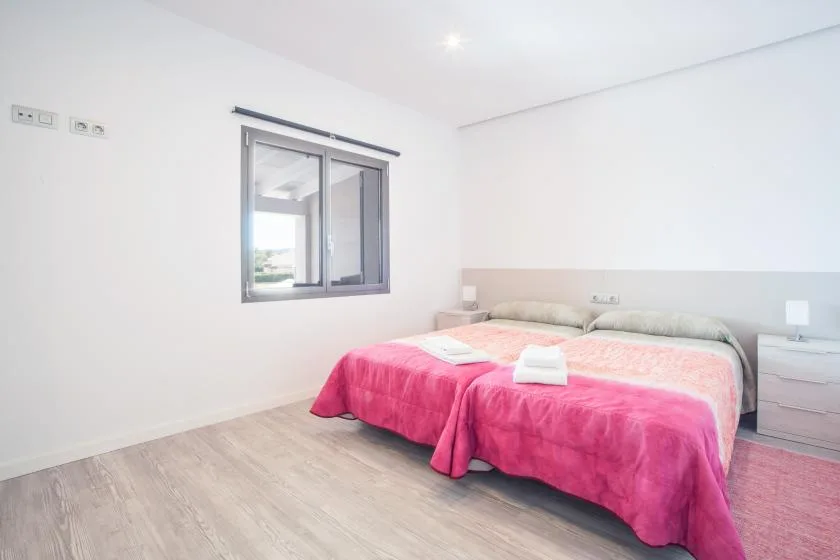 A bright bedroom with a double bed covered in a pink blanket beside a window.