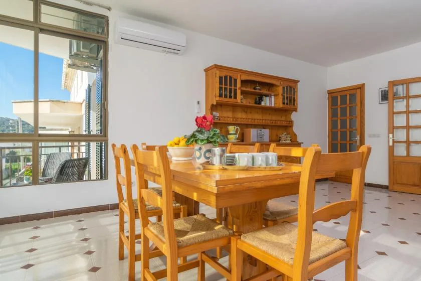 A wooden dining table and chairs sit in a bright room beside large windows.
