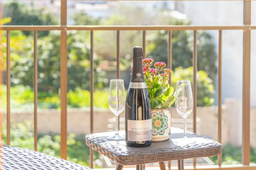 A balcony table holds a bottle, two glasses, and a small flowering plant.
