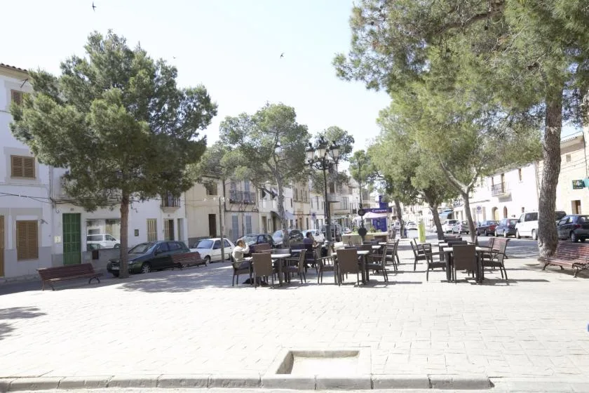 A sunny pedestrian square with outdoor tables and seating beneath leafy trees.
