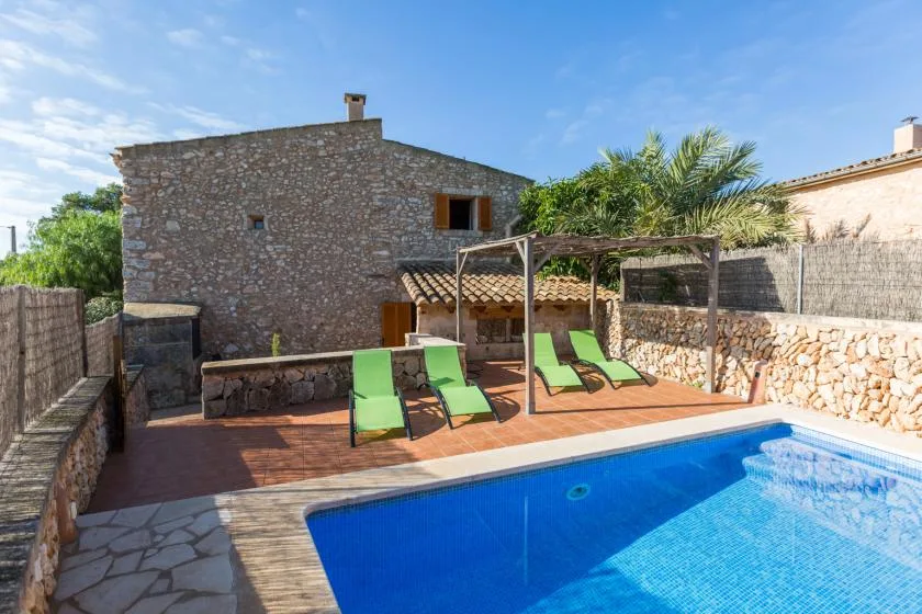 An outdoor swimming pool sits beside a stone house and a wooden deck with lounge chairs.