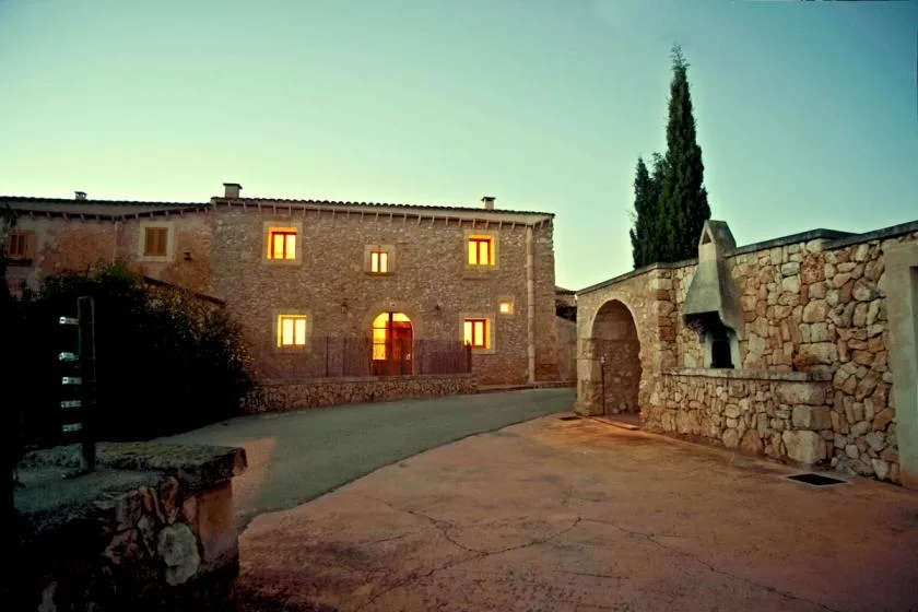 A stone building and arched stone wall face a paved courtyard at dusk.