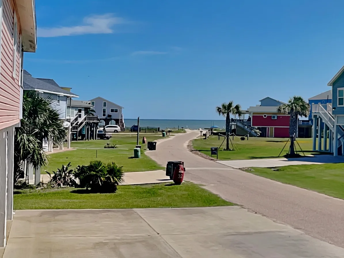 A paved residential street curves toward the ocean between houses and grassy lawns.