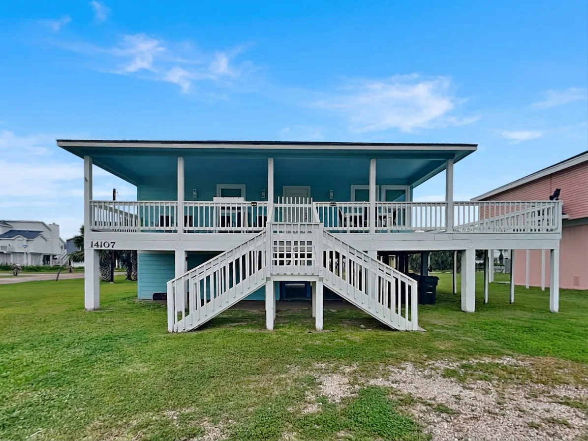 A raised coastal-style house with a broad covered deck and central stairs stands on a grassy lot.