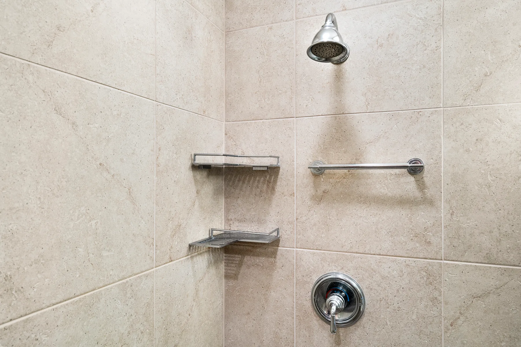 Una ducha con azulejos, cabezal, controles y estantes metálicos.