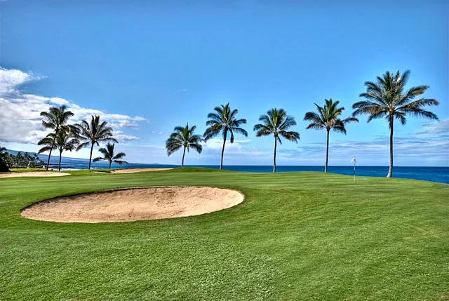 Un campo de golf costero con un búnker de arena, palmeras y vistas al océano.