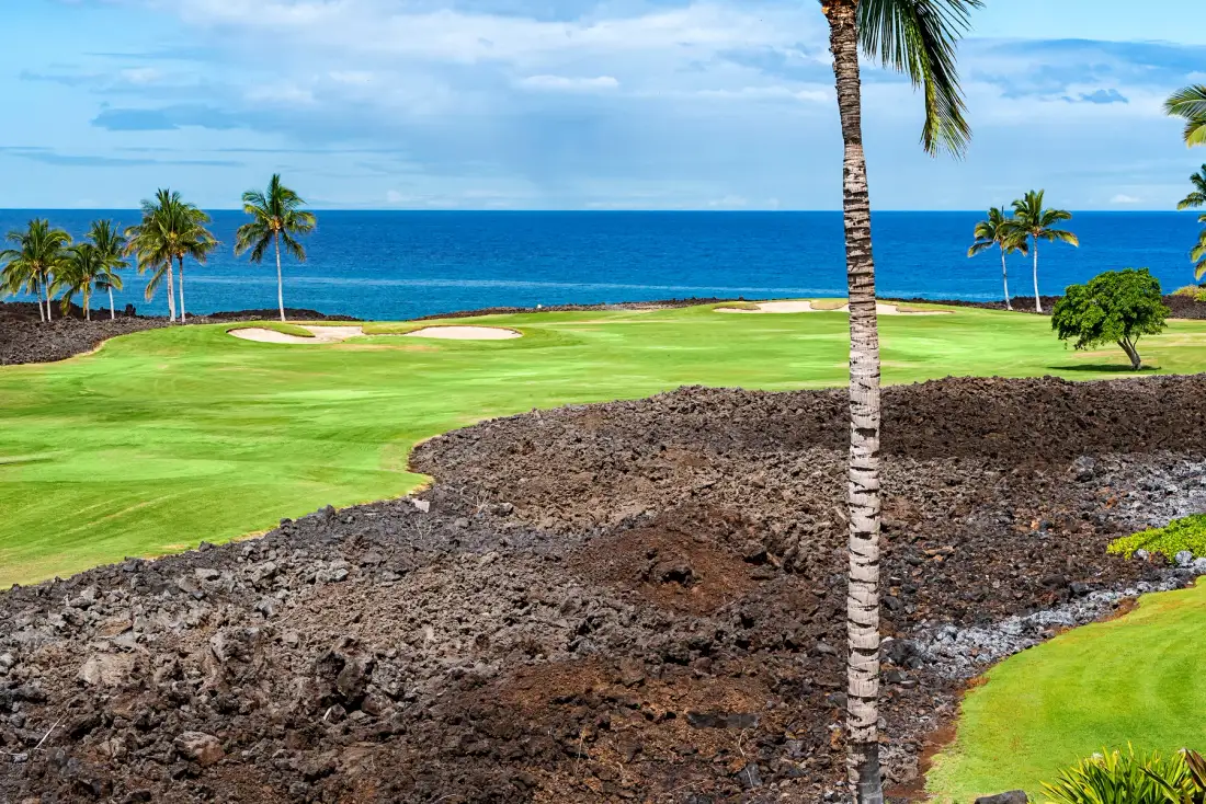 Un campo de golf costero con calles verdes, palmeras y vistas al océano.