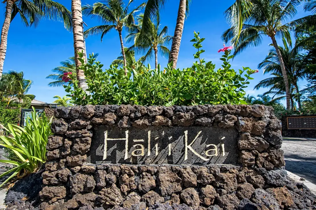 Un letrero de piedra que dice «Haliʻi Kai» está rodeado de plantas tropicales y palmeras.