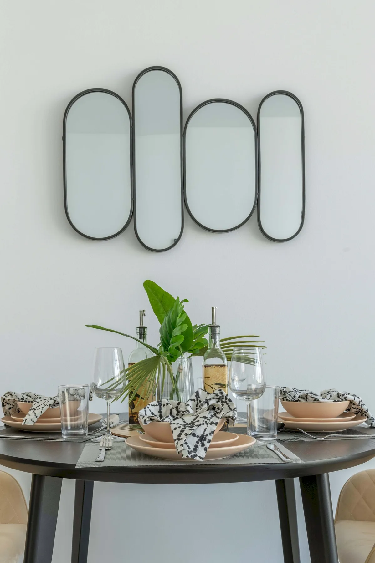 A dining table is set with plates, glasses, and a leafy centerpiece beneath a wall mirror.