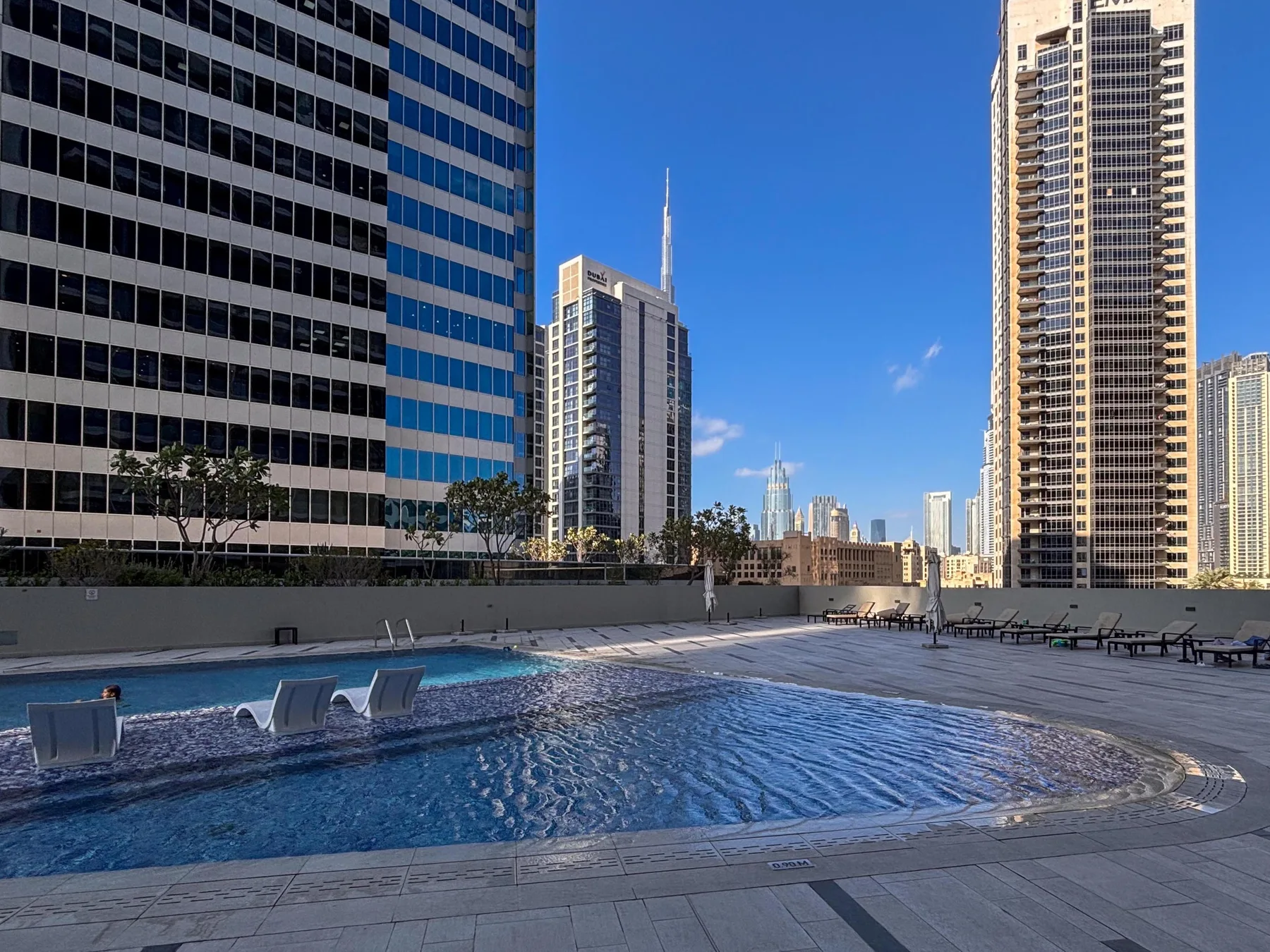A rooftop swimming pool sits among tall city buildings under a clear blue sky.