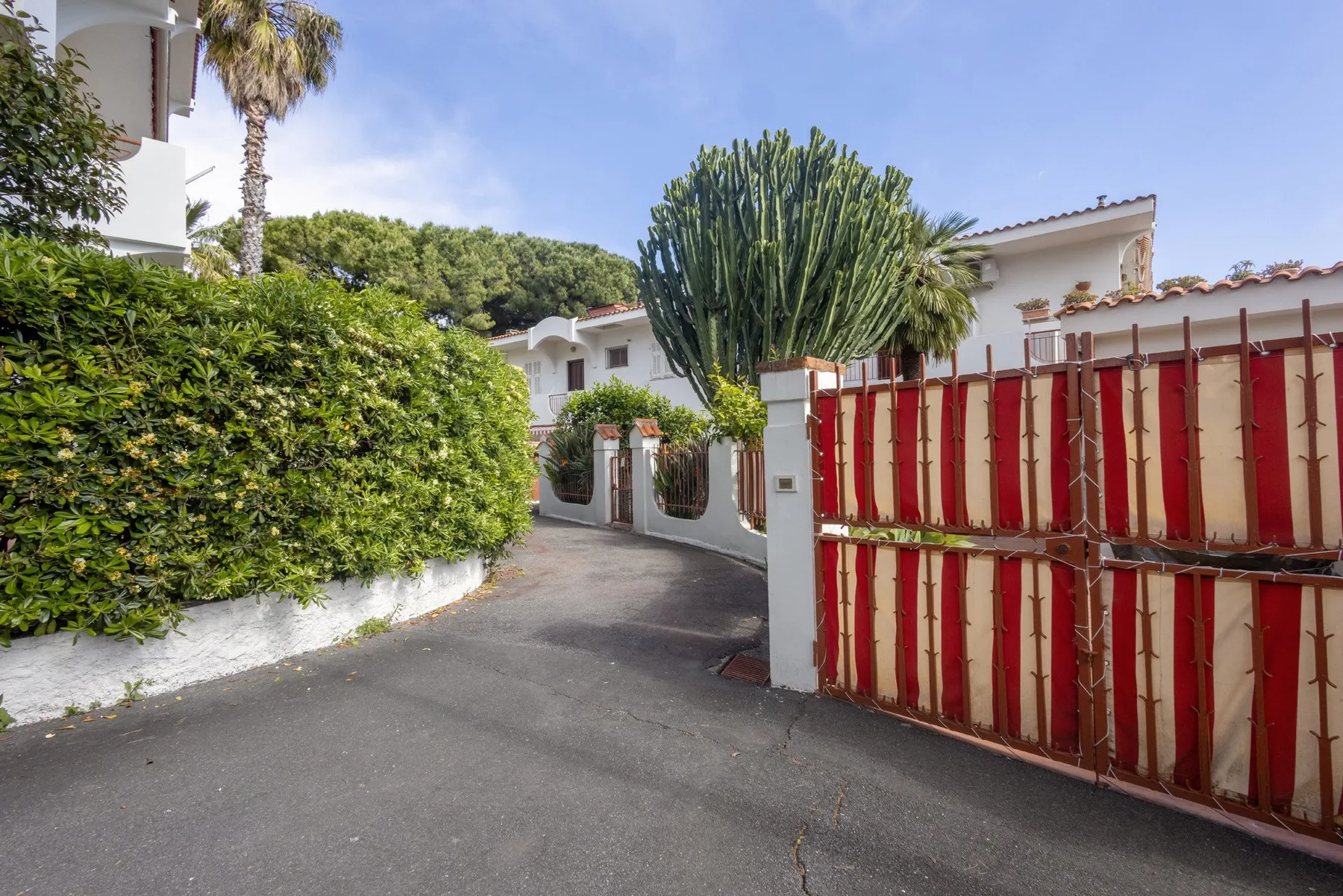 A paved driveway runs between white buildings, hedges, and a red gate.