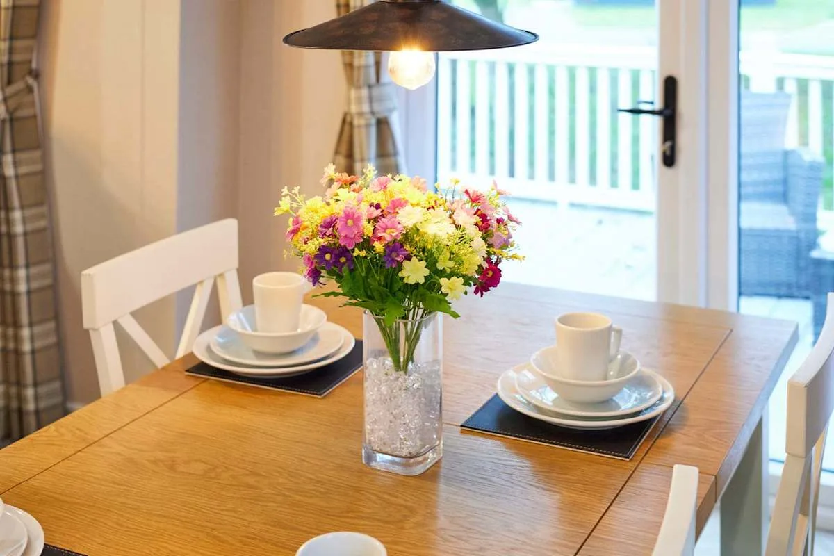 Una mesa de comedor está puesta con tazas y platos junto a un jarrón de flores coloridas.
