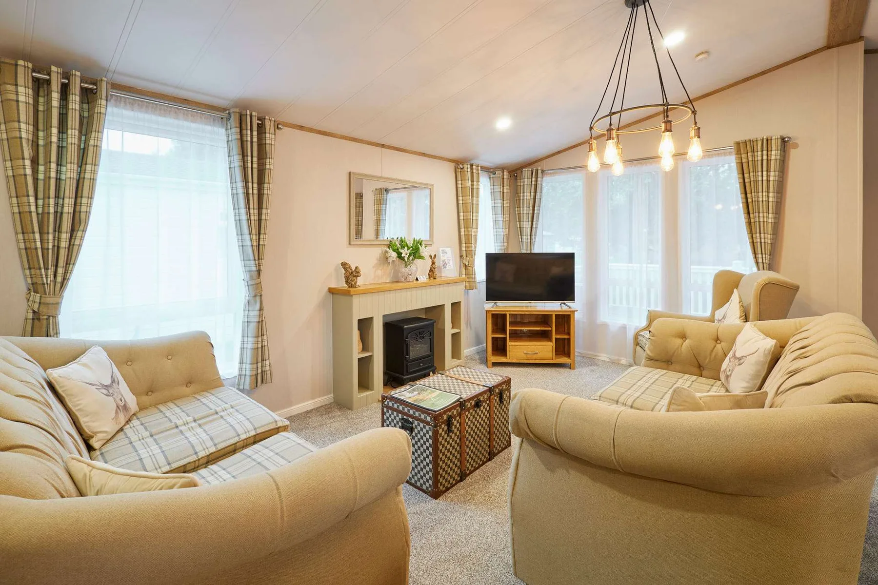 Una sala de estar con asientos beige, un televisor y grandes ventanas con cortinas.