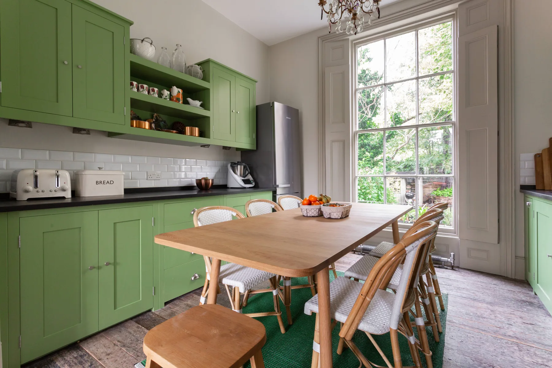 A dining table sits in a bright kitchen with green cabinets and a large window.