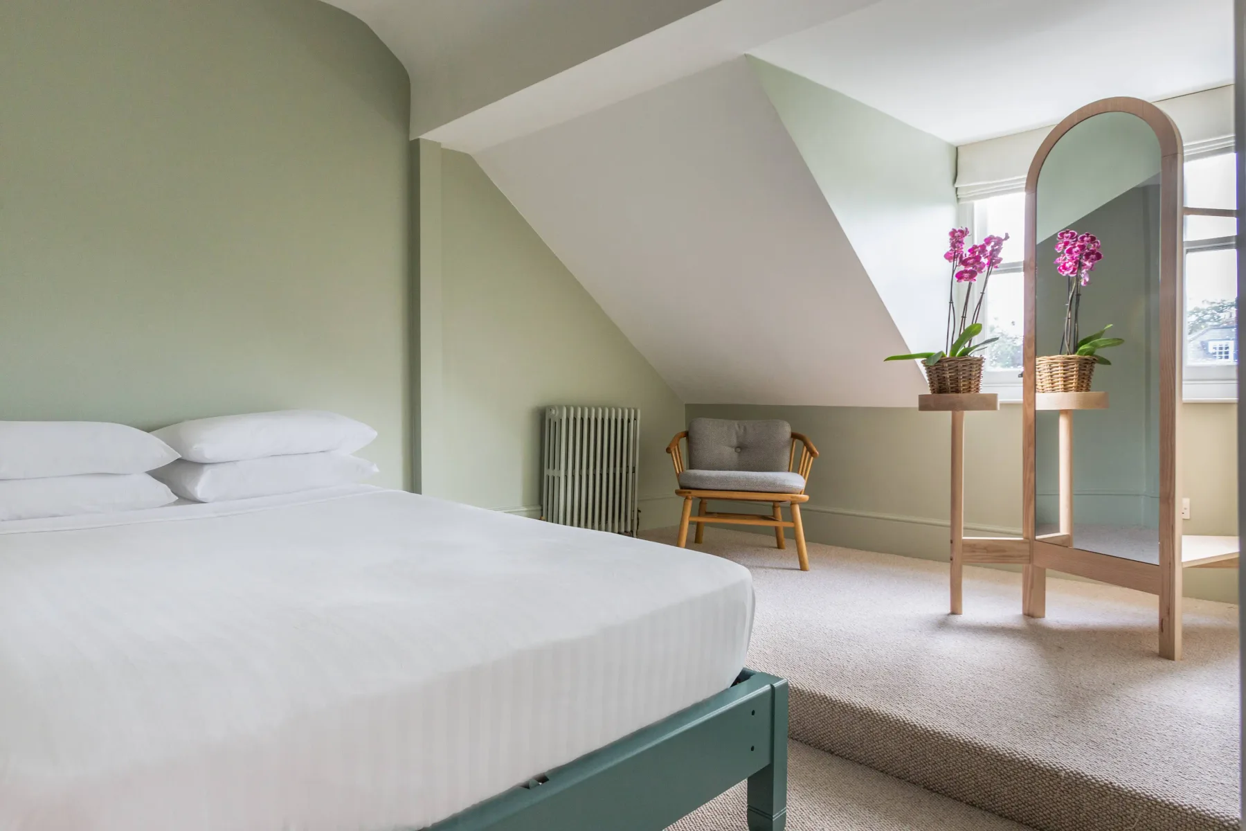 A bright bedroom with a bed, a chair, and a standing mirror.