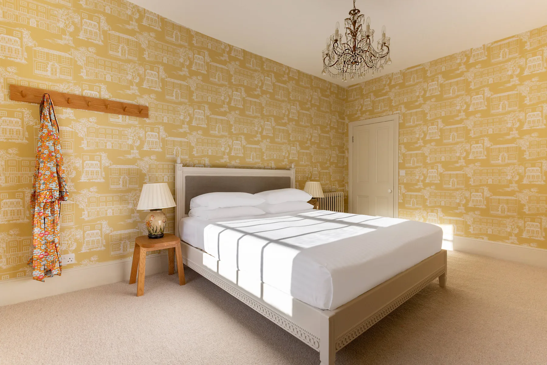 A bedroom with a neatly made bed, patterned yellow wallpaper, and a chandelier.