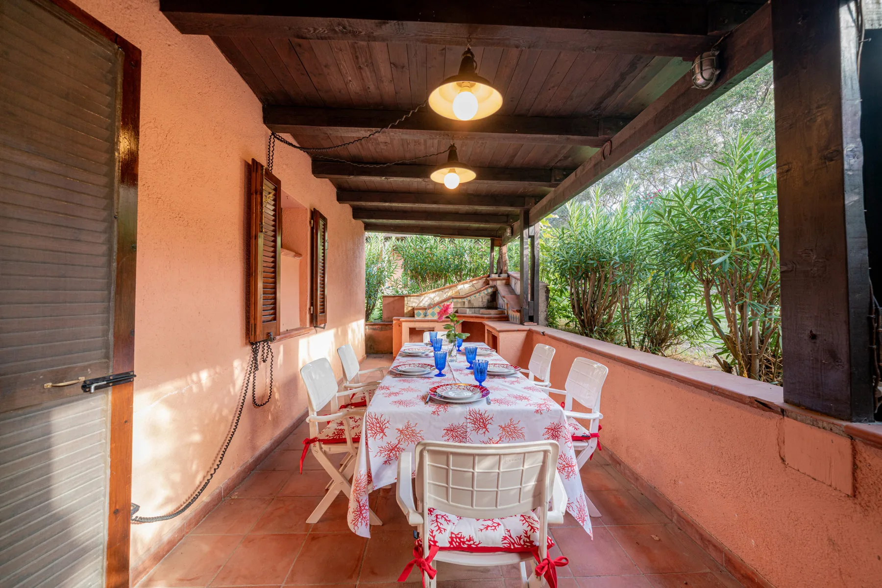 A covered outdoor dining area has a long table set beside lush greenery.