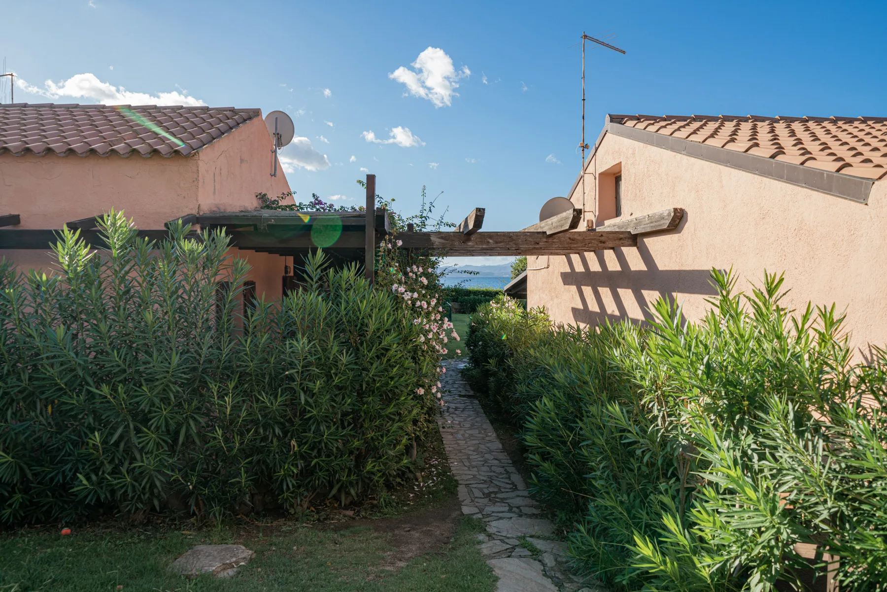 A stone path runs between leafy hedges toward two pale buildings beneath a blue sky.