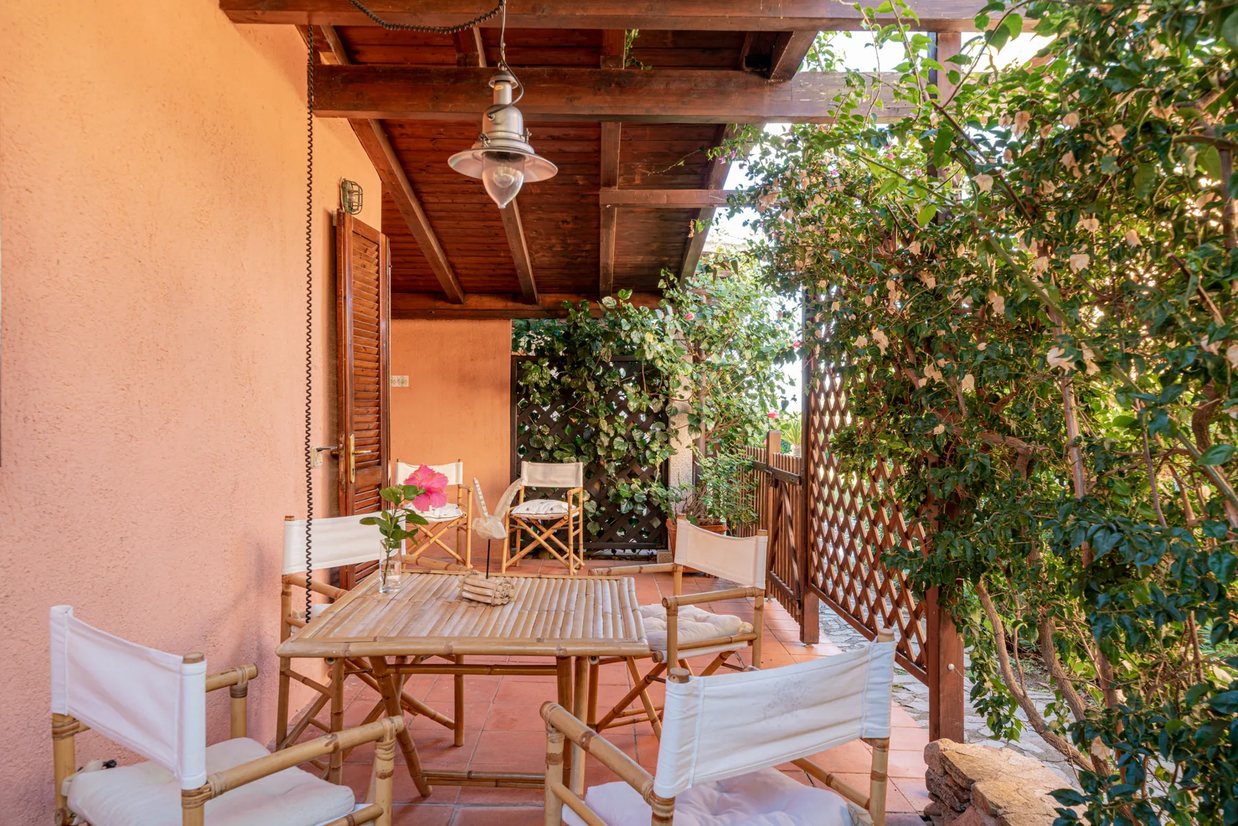 A covered outdoor patio has a wooden table and seating beside leafy greenery.