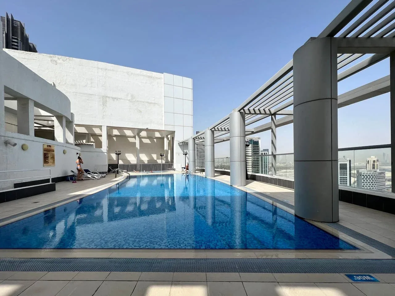 Una piscina en la azotea está junto a una terraza sombreada, con edificios de la ciudad a lo lejos.