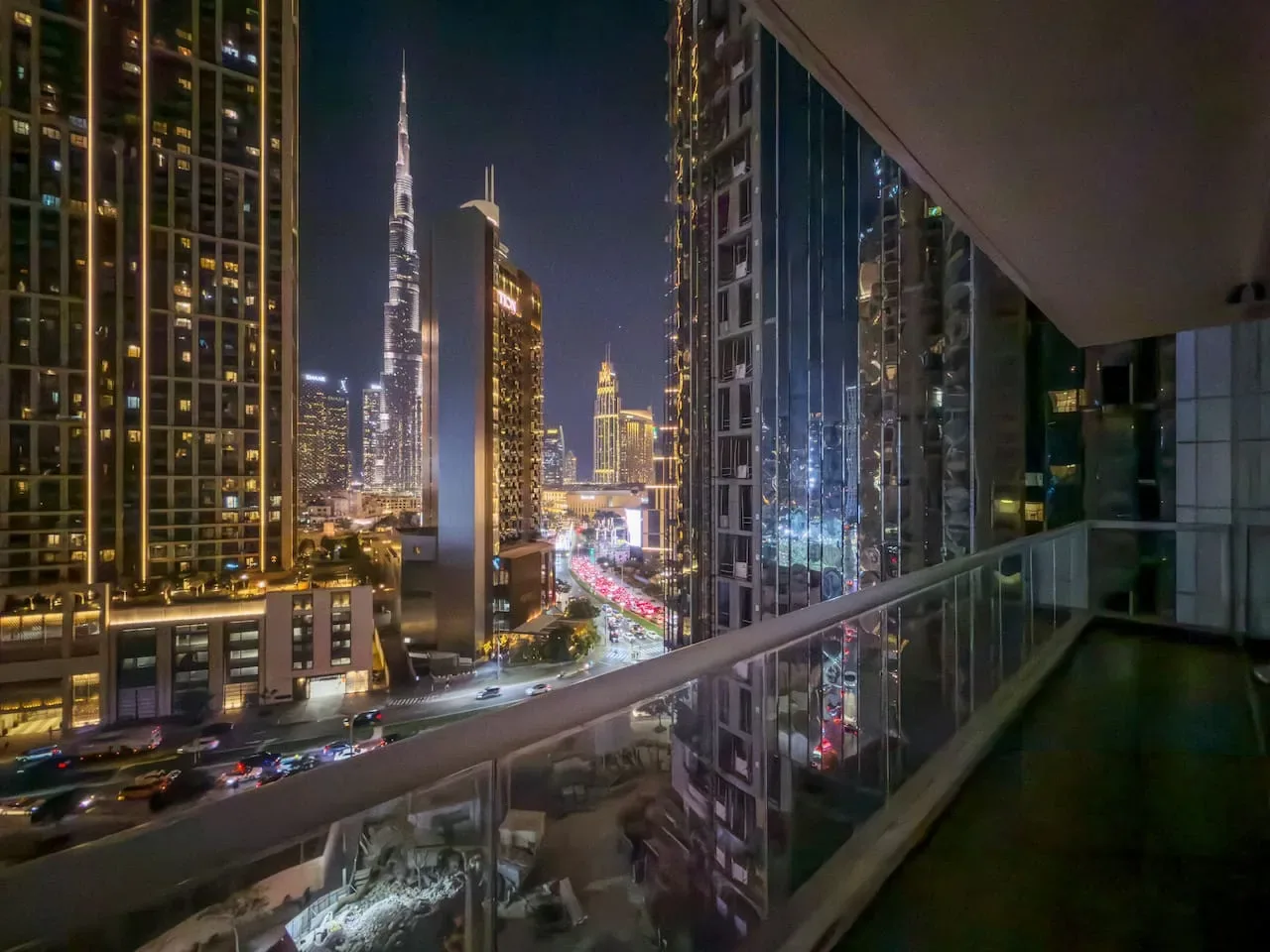 Un horizonte urbano nocturno se ve desde un balcón acristalado, con edificios iluminados y tráfico.