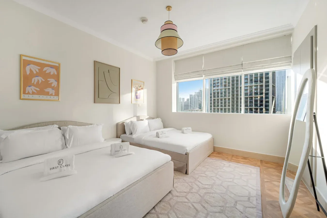 Un dormitorio luminoso con dos camas junto a una ventana amplia con vistas a edificios de la ciudad.