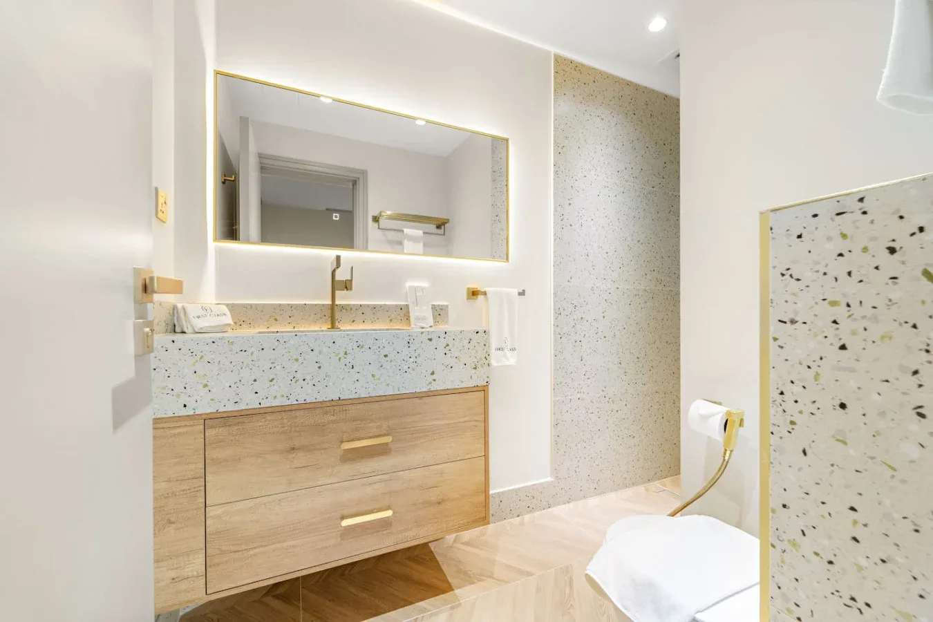 Un baño luminoso con mueble de madera, espejo iluminado y paredes moteadas.