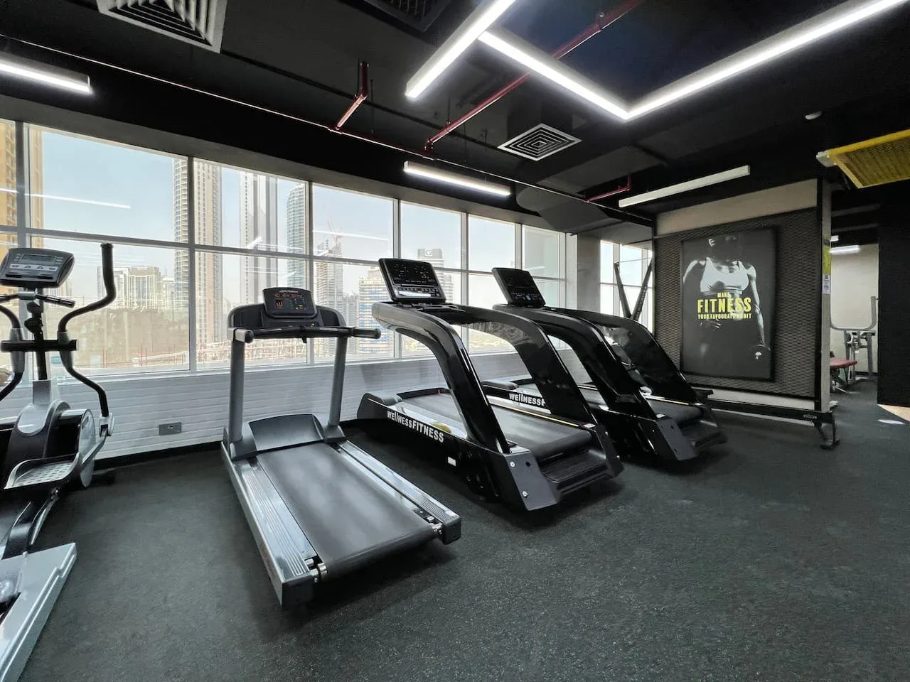 Un gimnasio con varias cintas de correr junto a grandes ventanas.