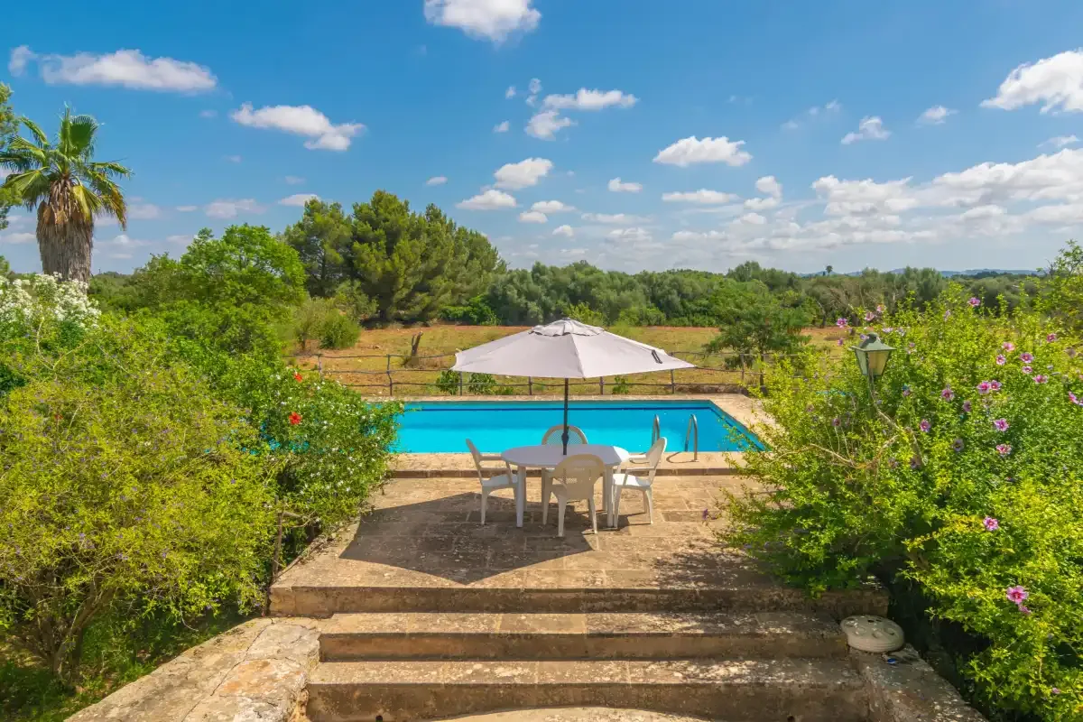 A garden patio with outdoor seating and an umbrella overlooks a swimming pool.