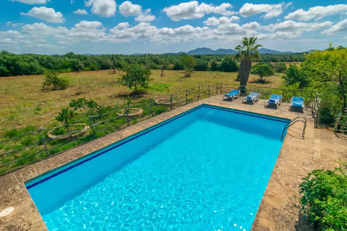 A bright outdoor swimming pool is surrounded by open countryside and trees.