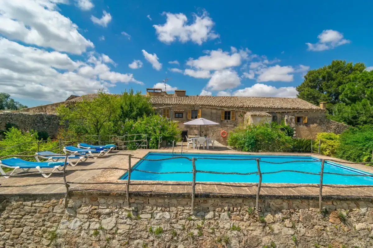 A swimming pool sits in front of a rustic house, surrounded by greenery.